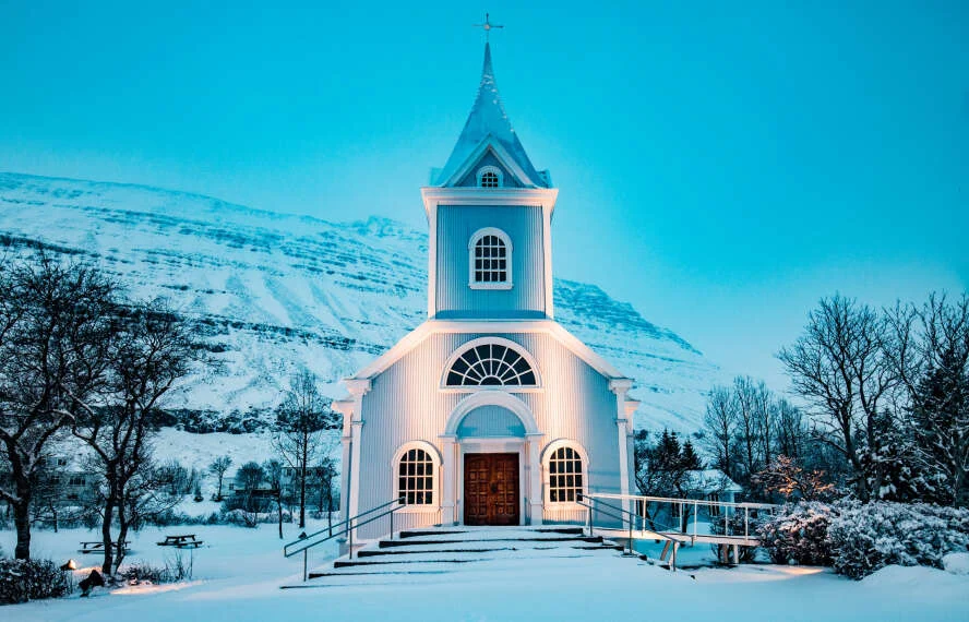 Pale blue church with a spire beneath snowy mountains